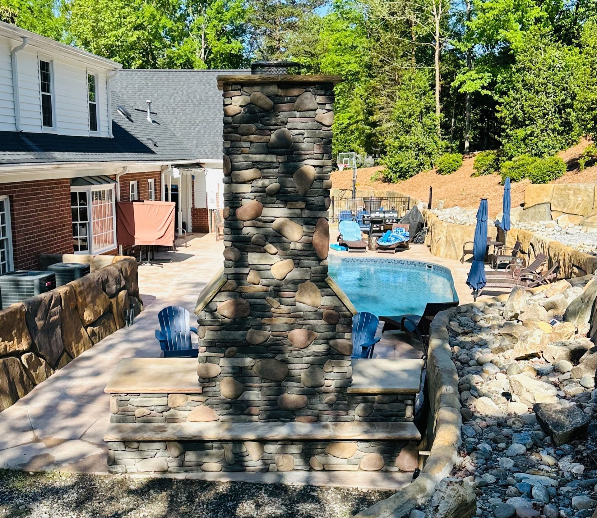 Backyard patio with a stone chimney, blue swimming pool, and stone retaining walls under trees