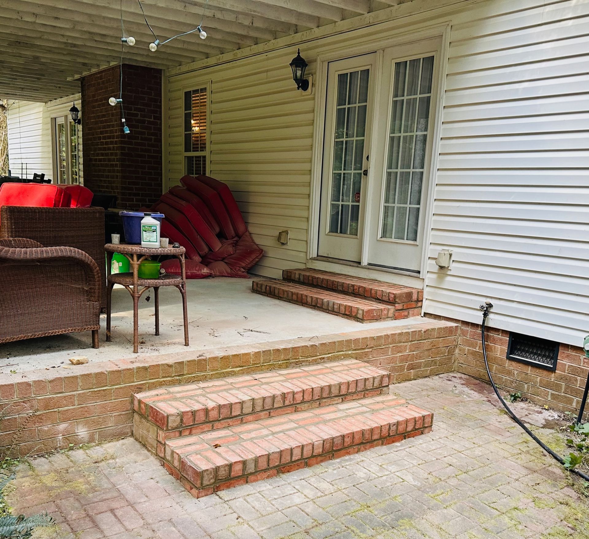 Covered porch with brick steps, white siding, wicker chair, and red cushions near a French door