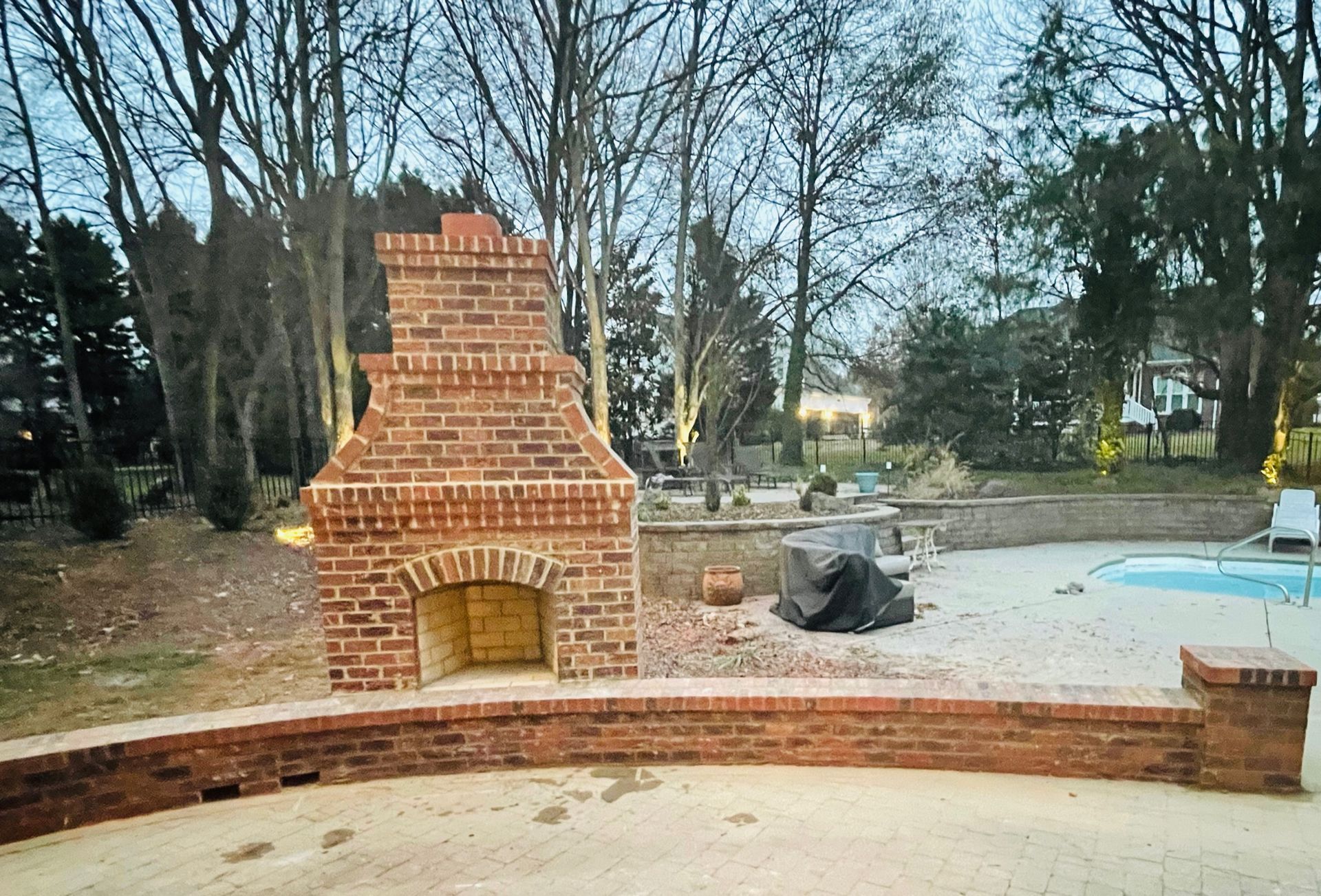 Outdoor brick fireplace in a backyard beside a pool and trees