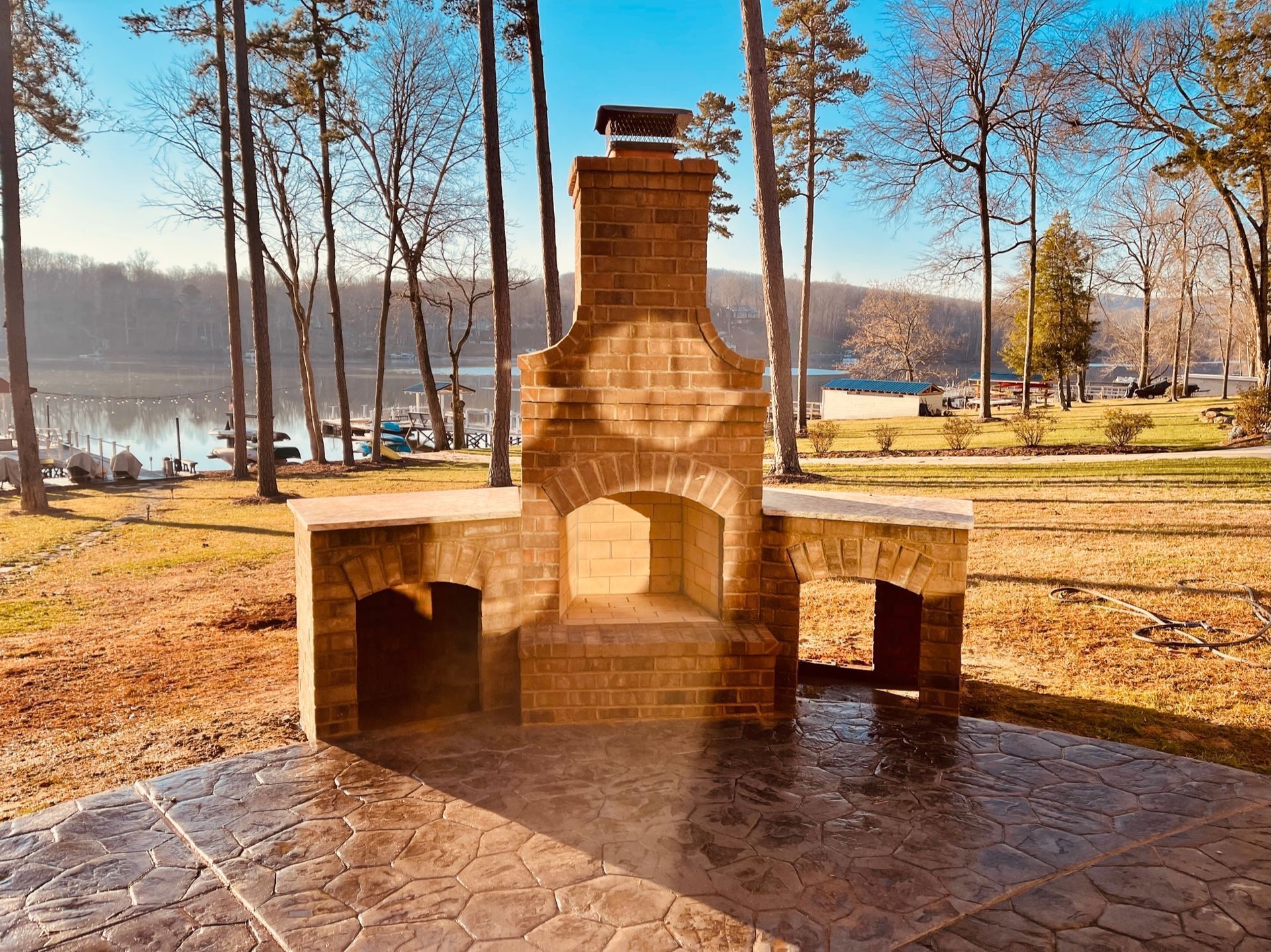 Stone outdoor fireplace with benches in a sunny park by a lake, surrounded by bare trees