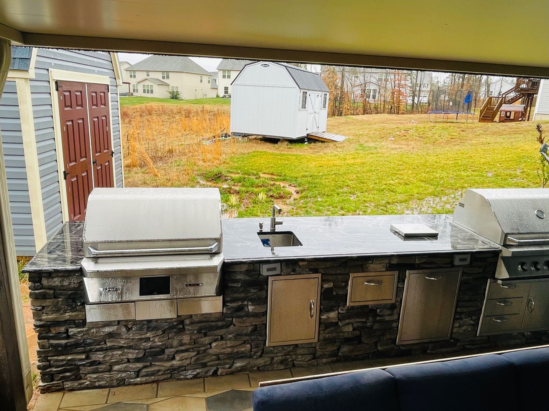 Outdoor stone patio with two stainless steel grills and a sink, overlooking a grassy yard and white shed.