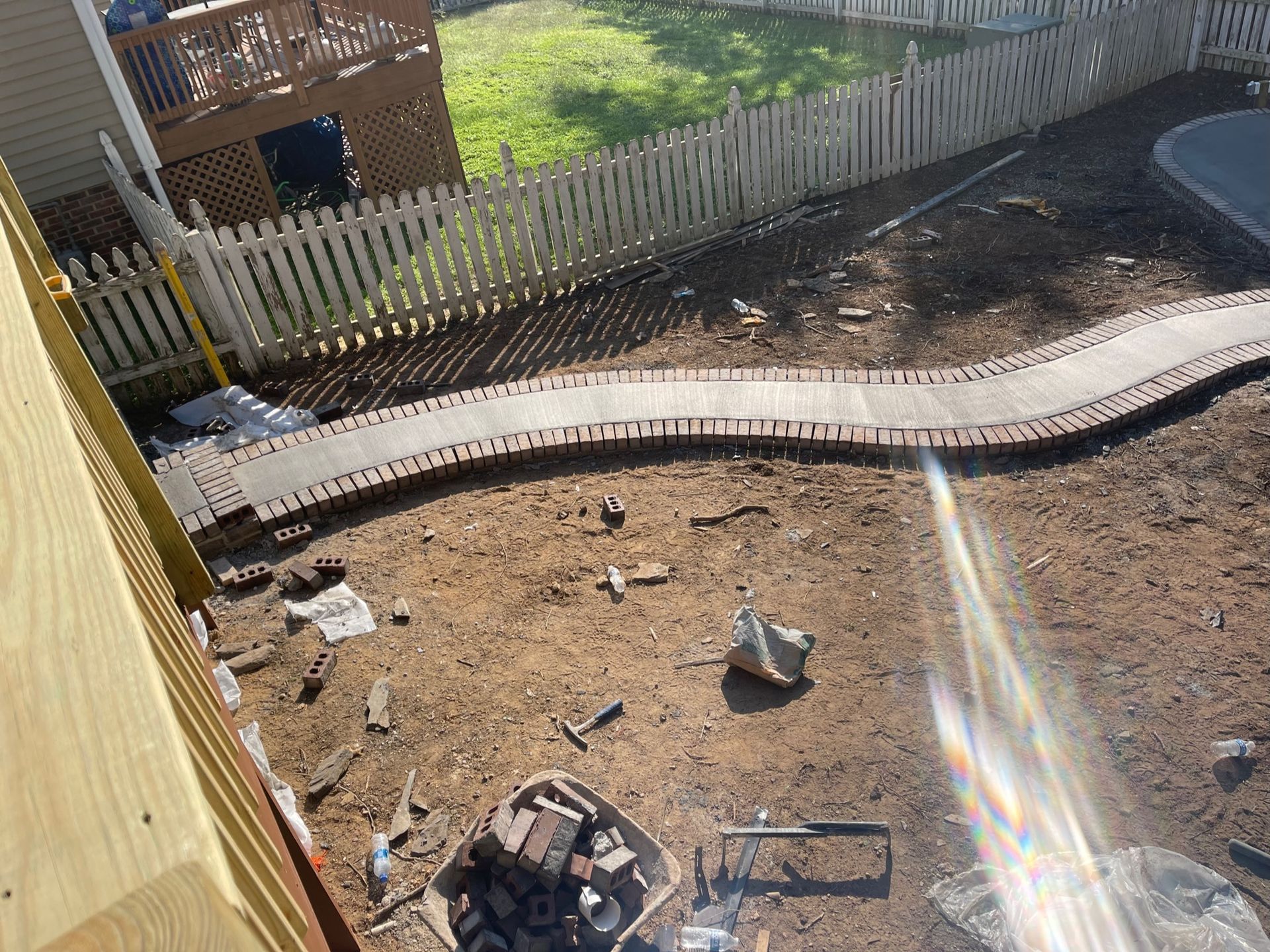 Backyard construction site with curved concrete edging, dirt, tools, and a white picket fence.