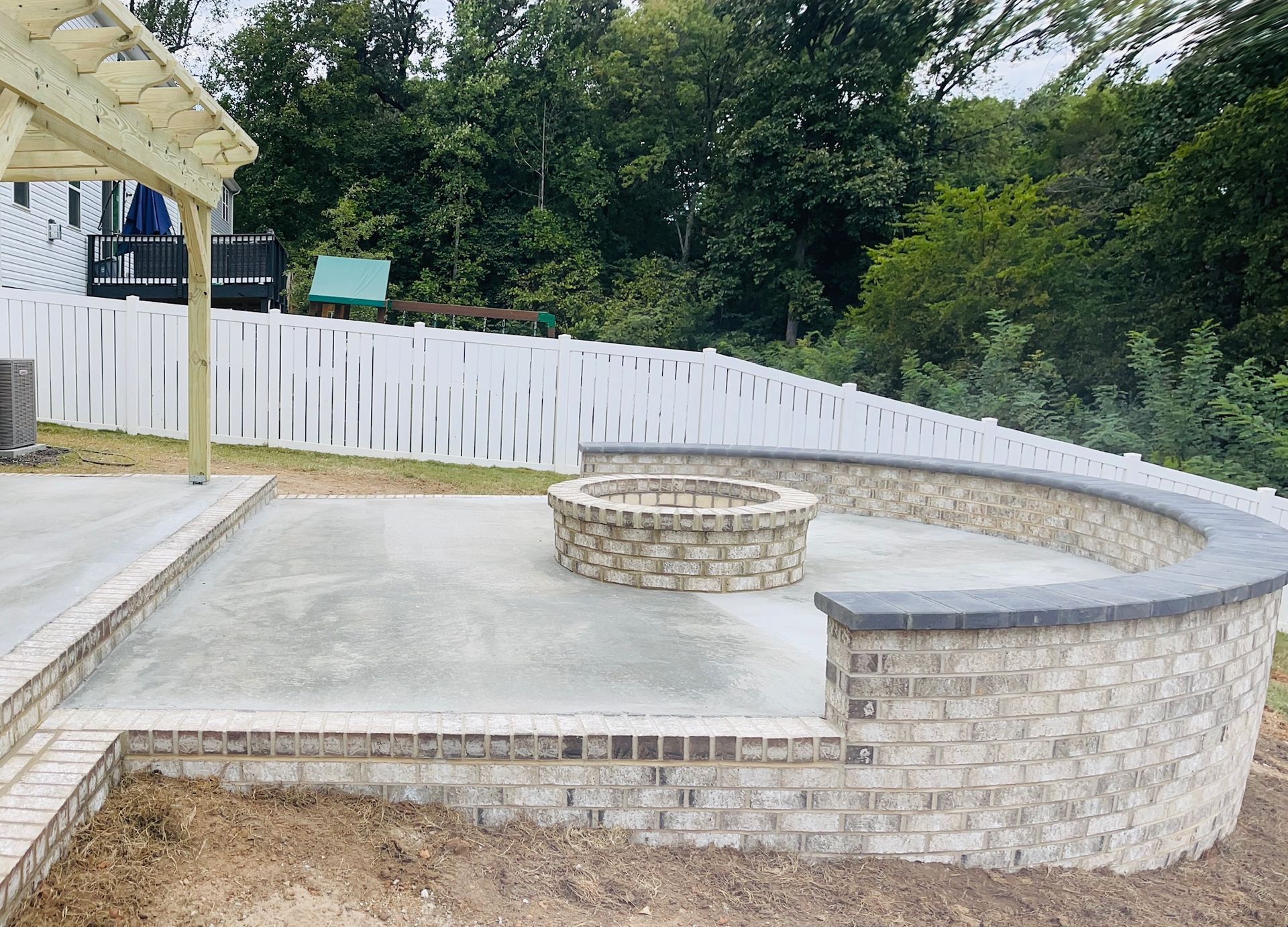 Concrete patio with curved stone seating wall and circular fire pit beside a white fence and trees