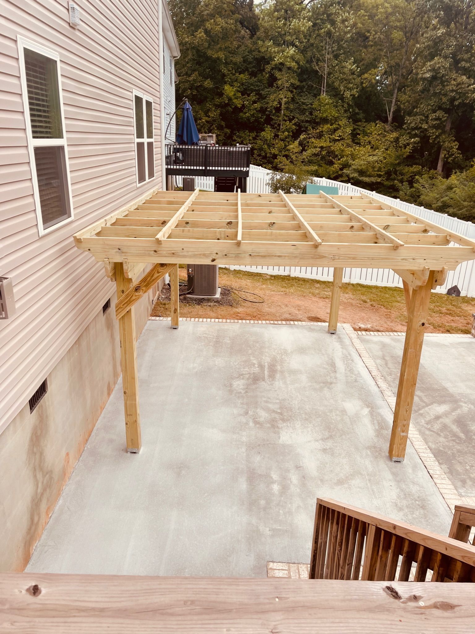 Wooden deck frame under construction beside a house, with support posts and joists over a concrete patio.