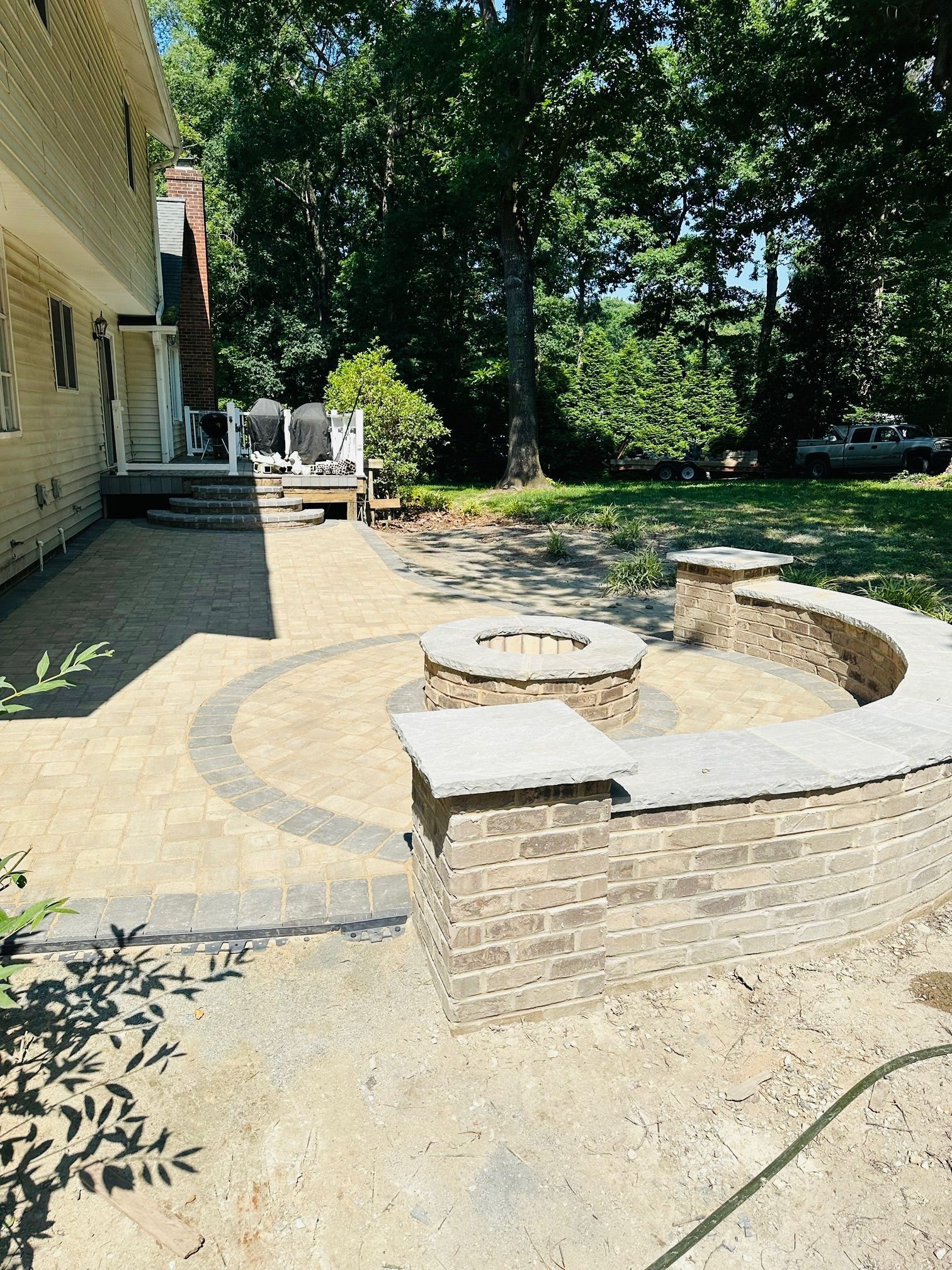 Backyard stone patio with curved brick fire pit beside a beige house and trees in shade