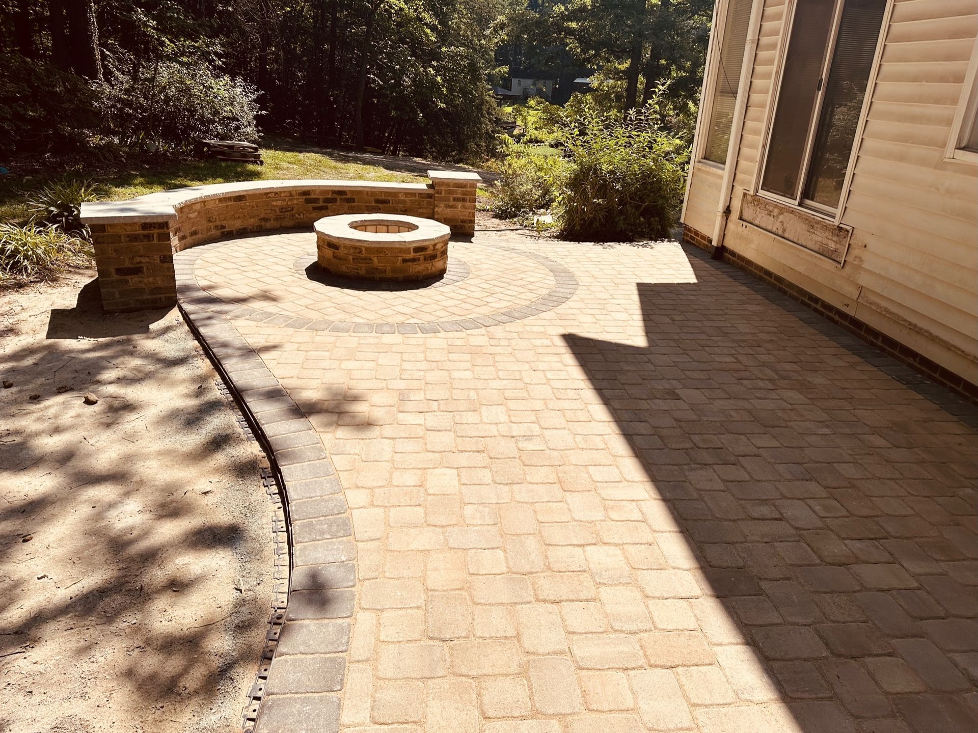 Brick patio with curved stone bench and fire pit beside a house, in bright sunlight.
