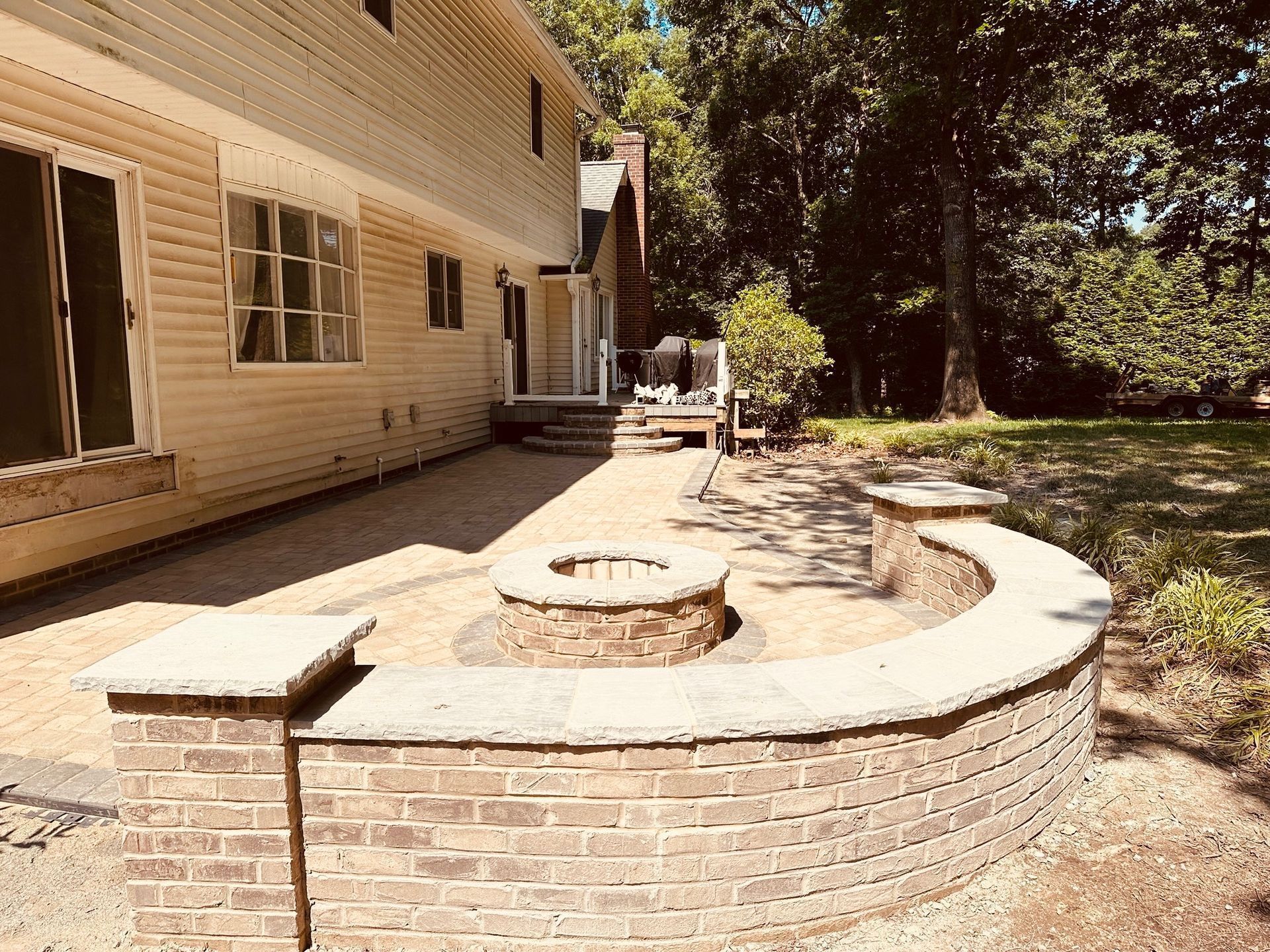Brick patio and curved retaining wall beside a beige house, with steps and trees in the yard.