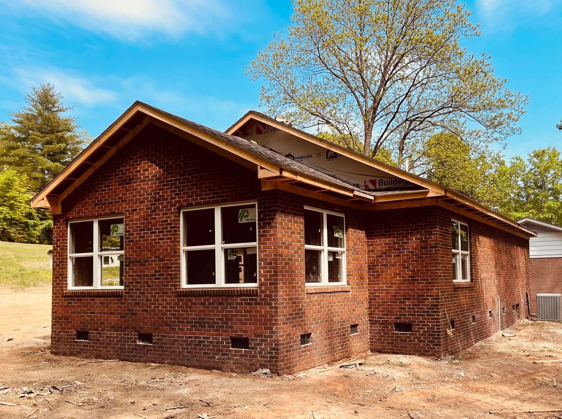 Brick house under construction with unfinished roof and window frames in a dirt yard