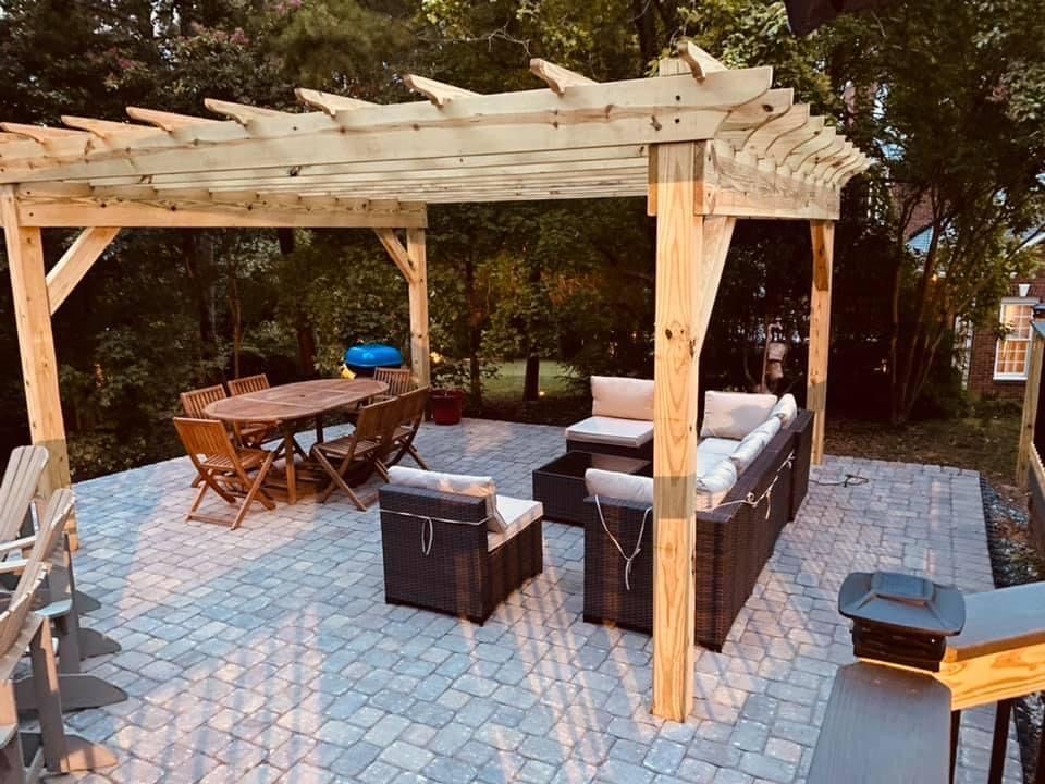 Backyard patio with a wooden pergola, cushioned seating, table, and paver flooring at dusk