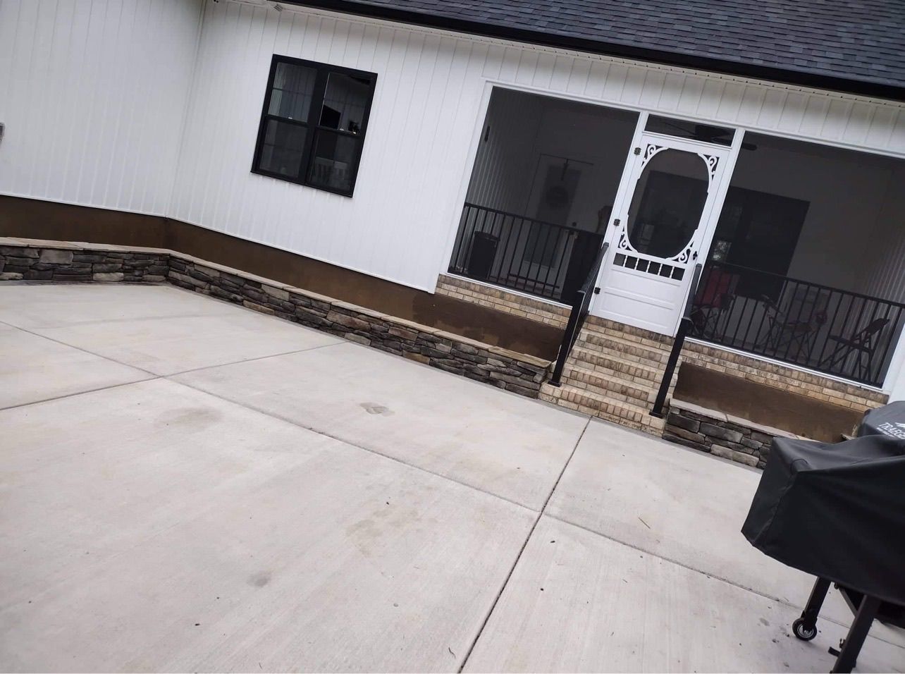 Outdoor concrete patio facing a white house with steps, a front door, and a covered grill on the right.