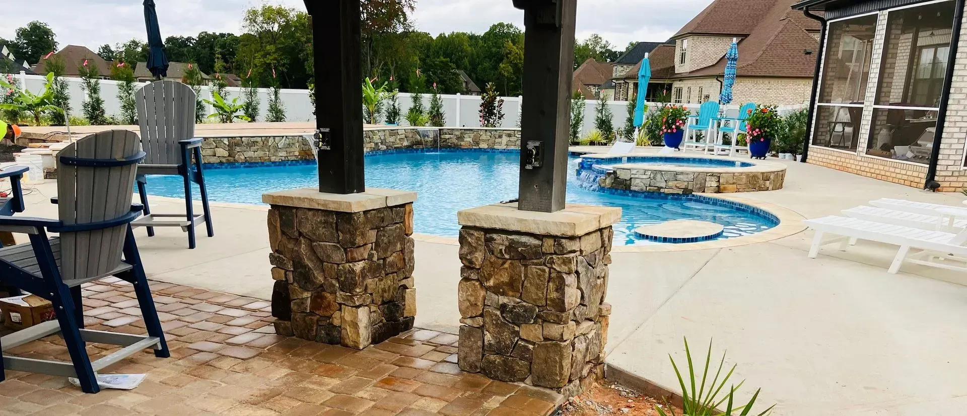 Backyard pool patio with lounge chairs, stone pillars, and a small hot tub under a covered pergola.