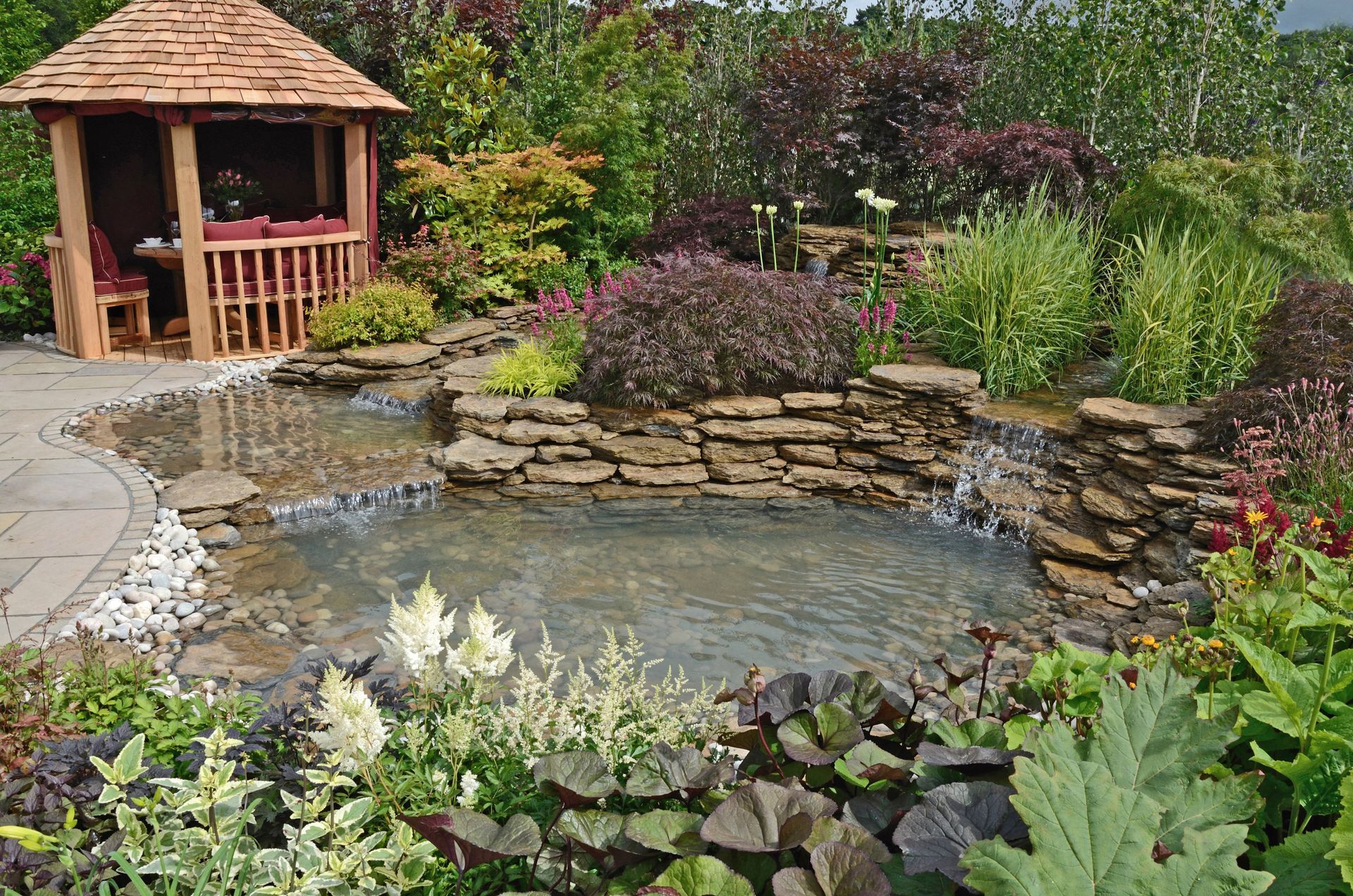 Garden pond with a small wooden gazebo, stone waterfall, and colorful flowers and greenery