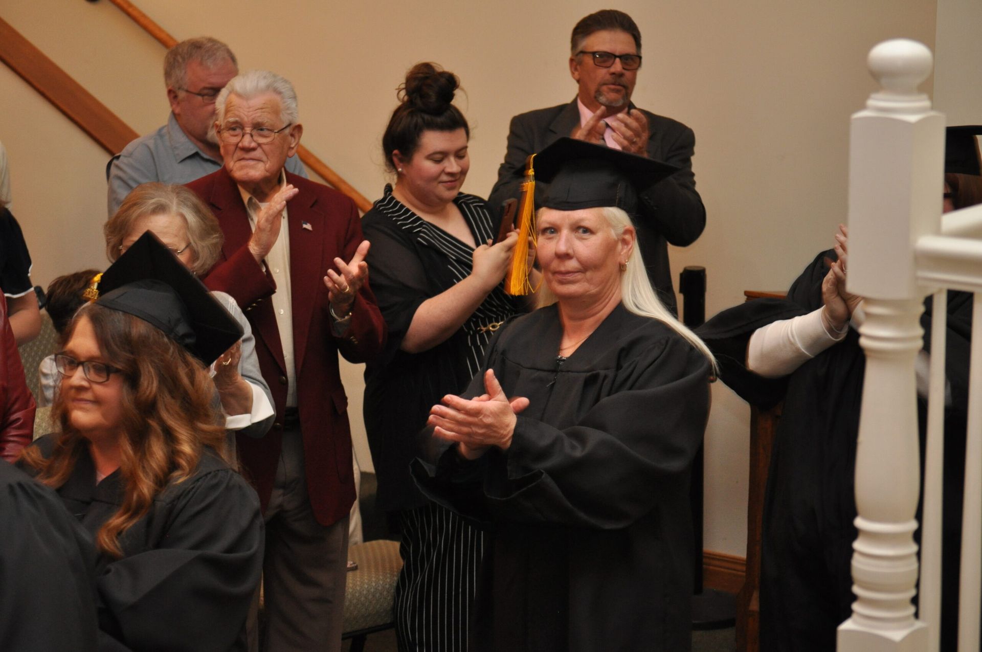 Graduates in caps and gowns clap, standing on stairs. People in the background applaud.