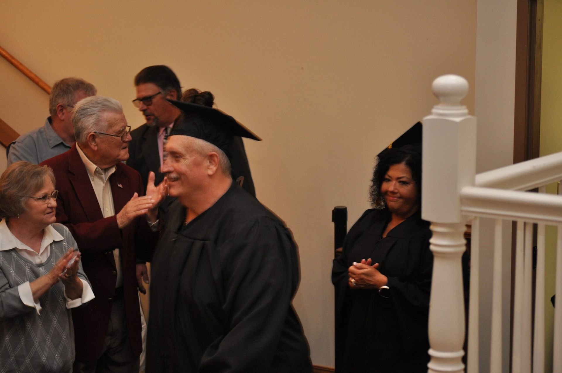 A man in a graduation gown smiles as people clap and celebrate on a staircase.