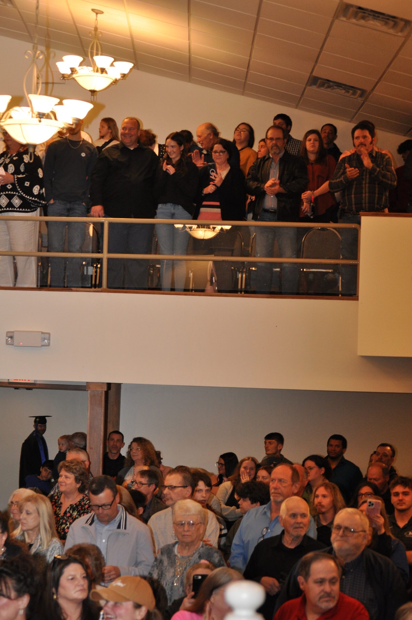 People gathered in a two-story building. Some are on a balcony, others below. Ceiling lights illuminate the crowd.