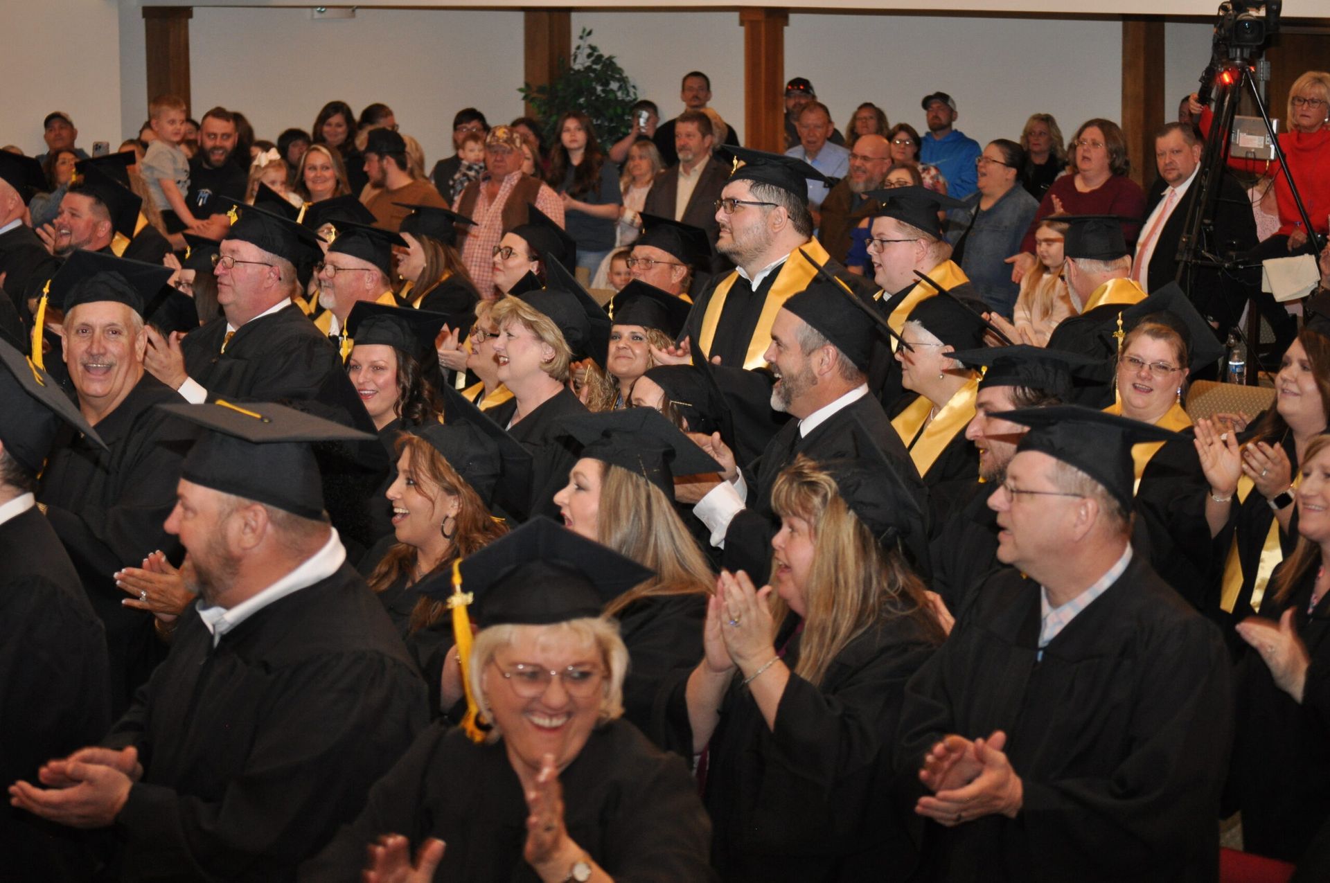 Graduation ceremony: graduates in black gowns and mortarboards applaud, large crowd in a hall.