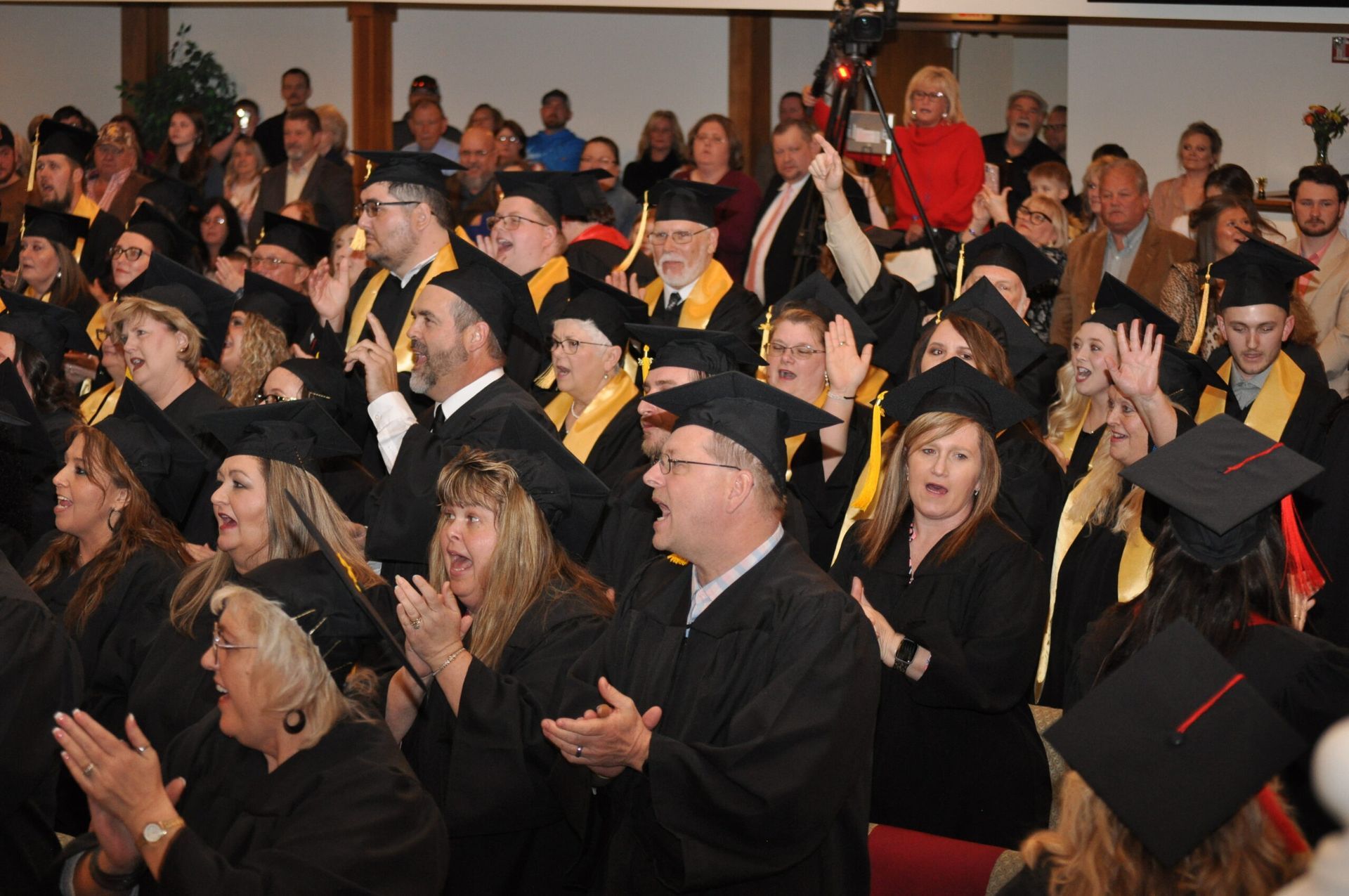 Graduation ceremony: graduates in black caps and gowns, clapping and cheering, in a crowded hall.