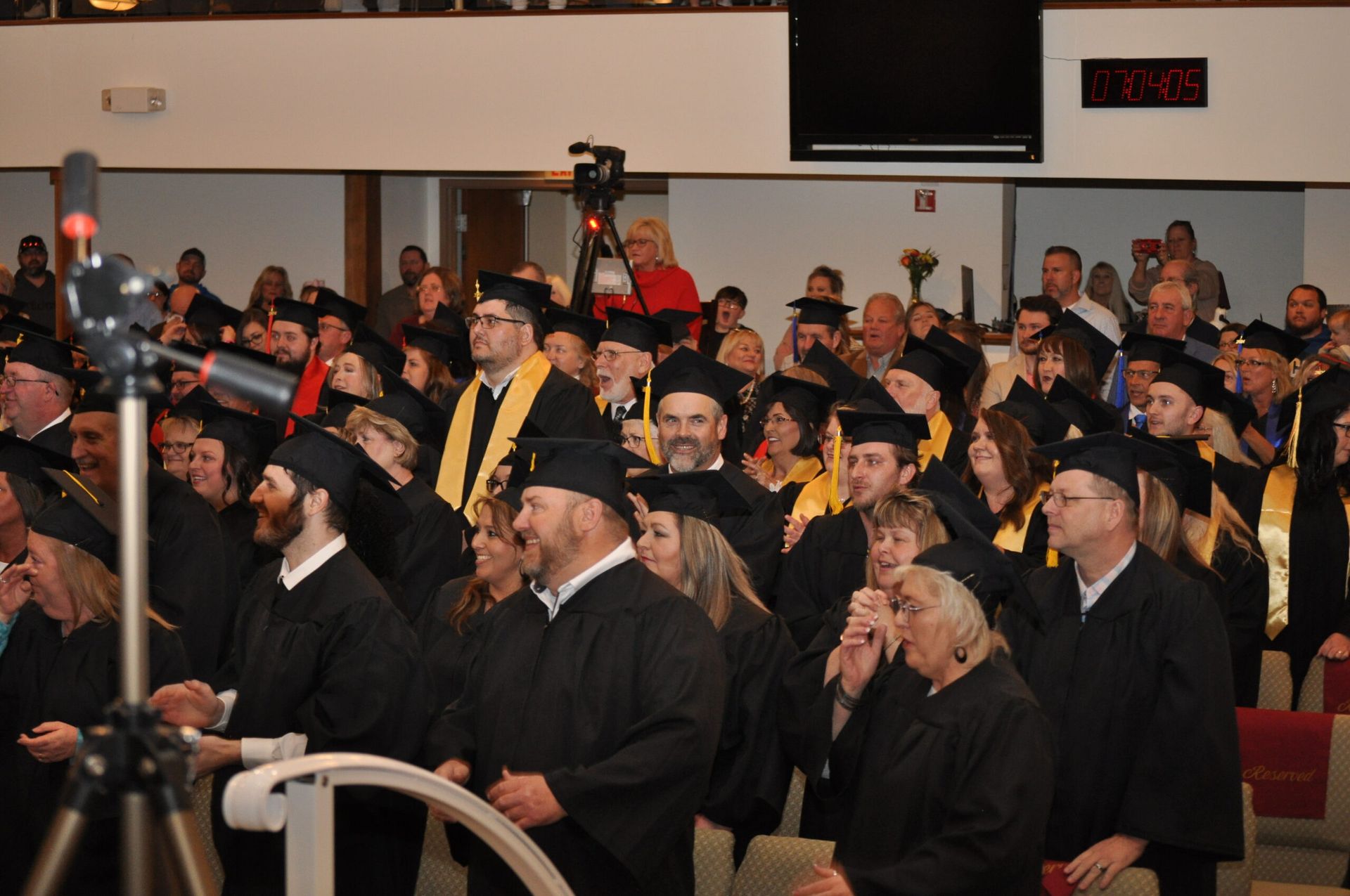 Graduation ceremony: graduates in black gowns and mortarboards inside a large hall, some with gold stoles.