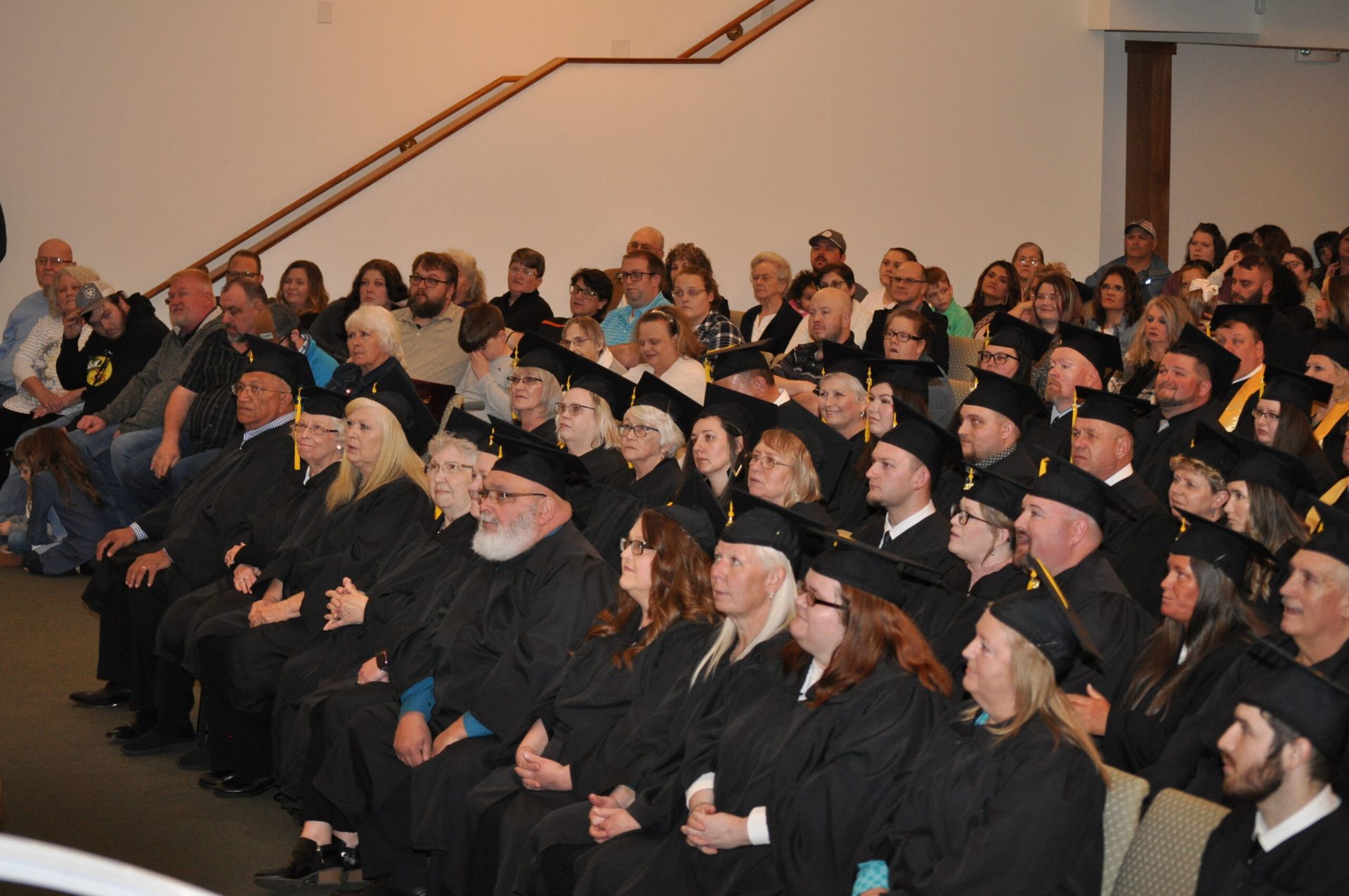 Graduation ceremony, graduates in black gowns and caps, seated audience, indoor setting.
