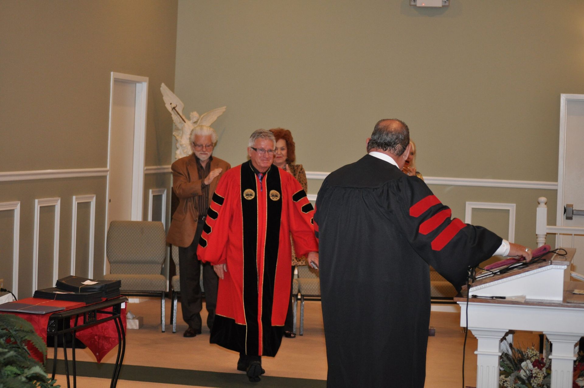 People in academic robes in a church setting. One in red and black robe walks forward. Others stand nearby.
