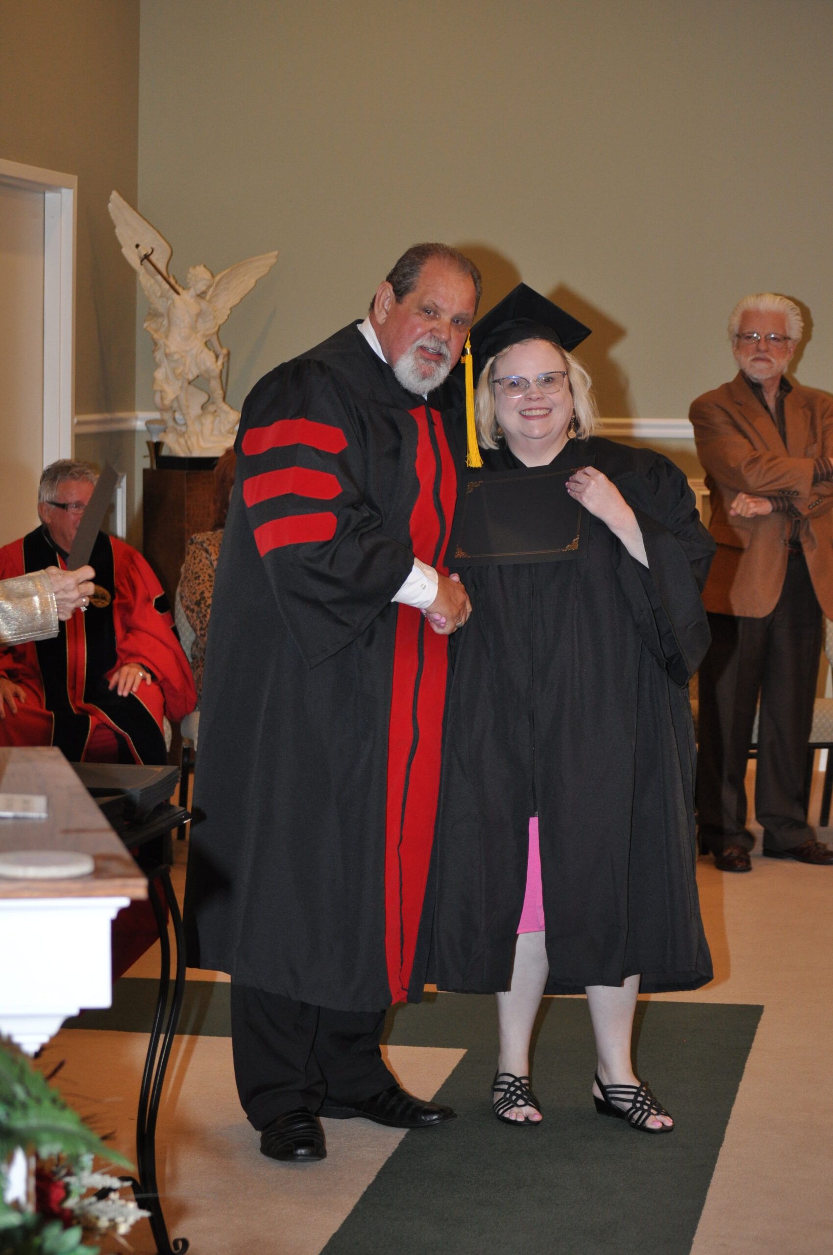 Man in robe shakes hands with graduate wearing cap and gown. Inside a room.
