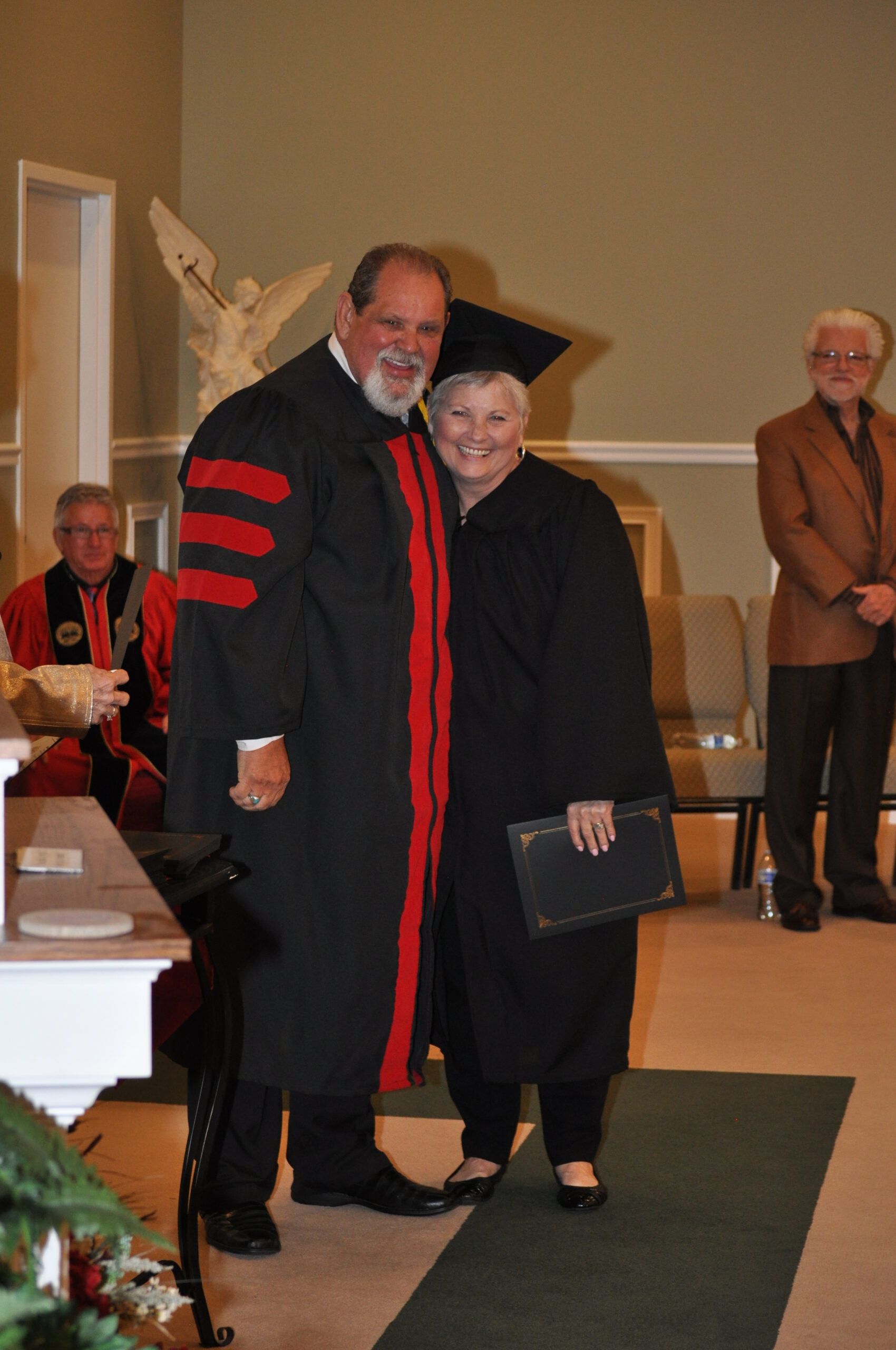 A graduate in cap and gown embraces a person in academic robes at a graduation ceremony.