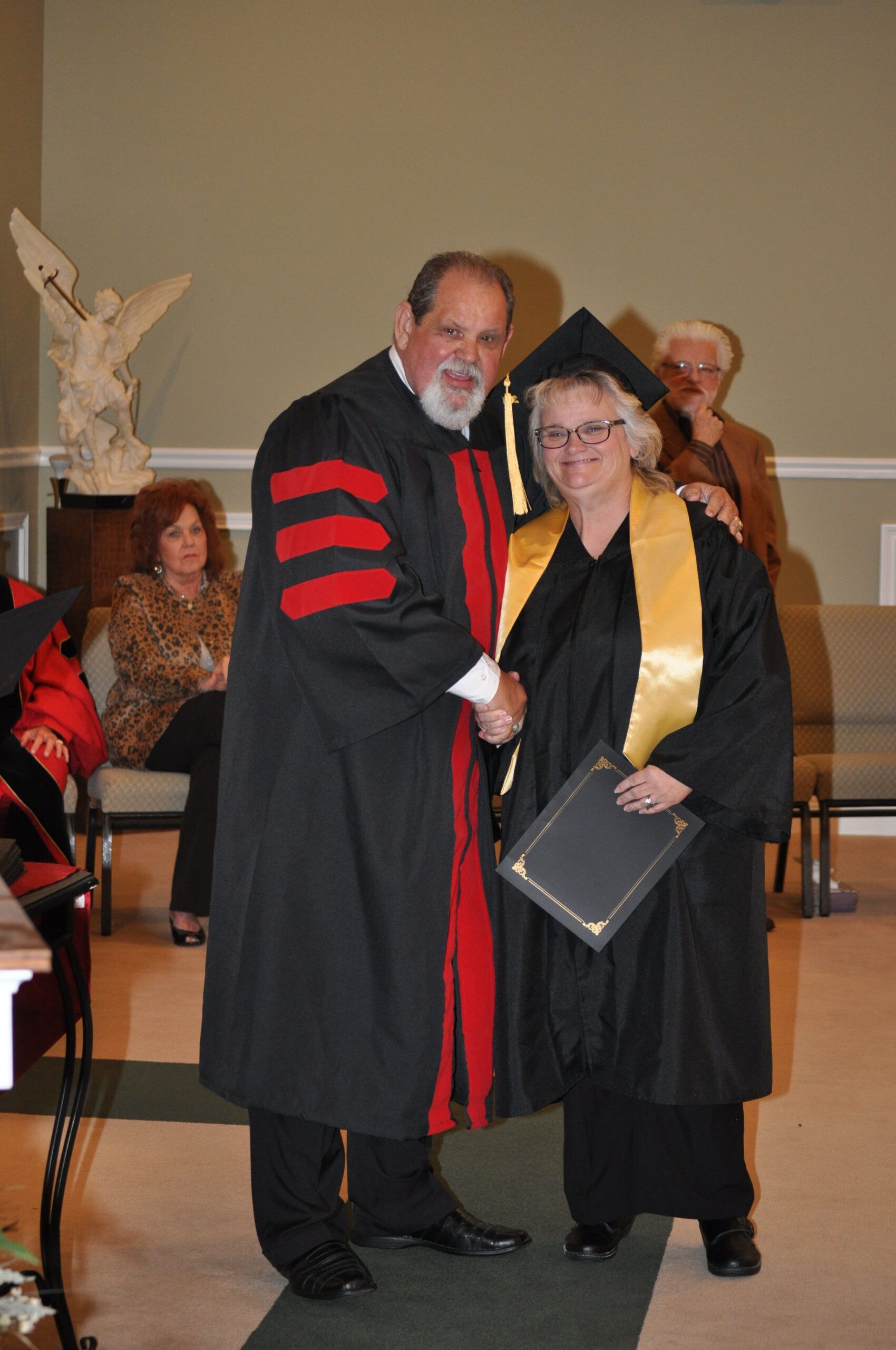 A person in graduation regalia shakes hands with another person in formal attire; indoor setting.