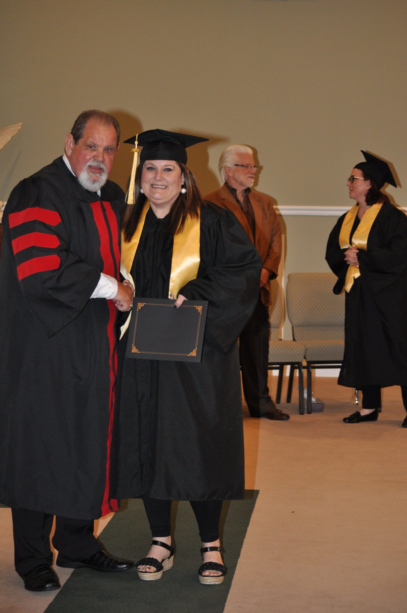 Person in graduation gown shaking hands with a robed figure at ceremony; others watch.