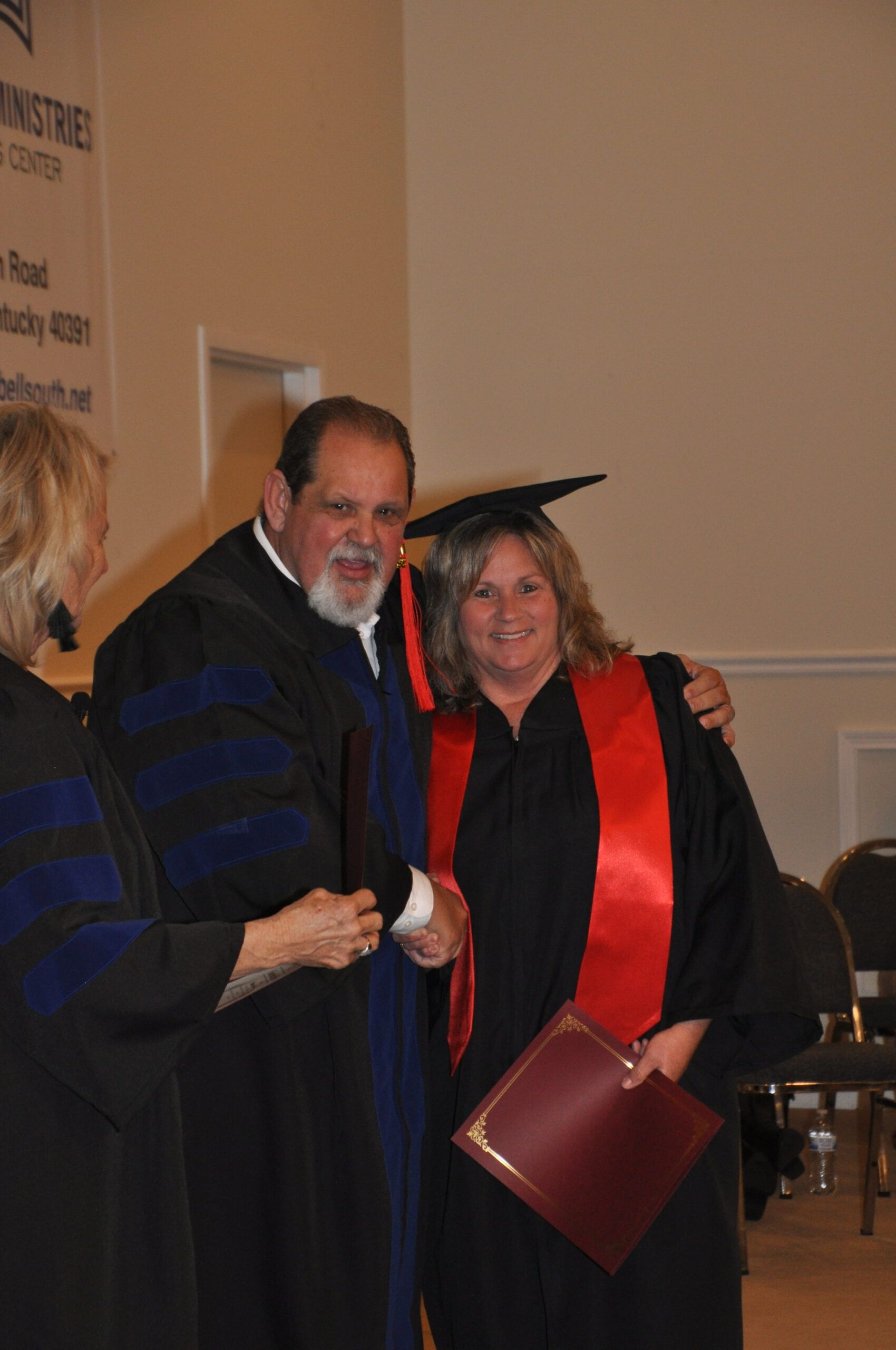 Man in academic robe shakes hands with graduate wearing red sash.