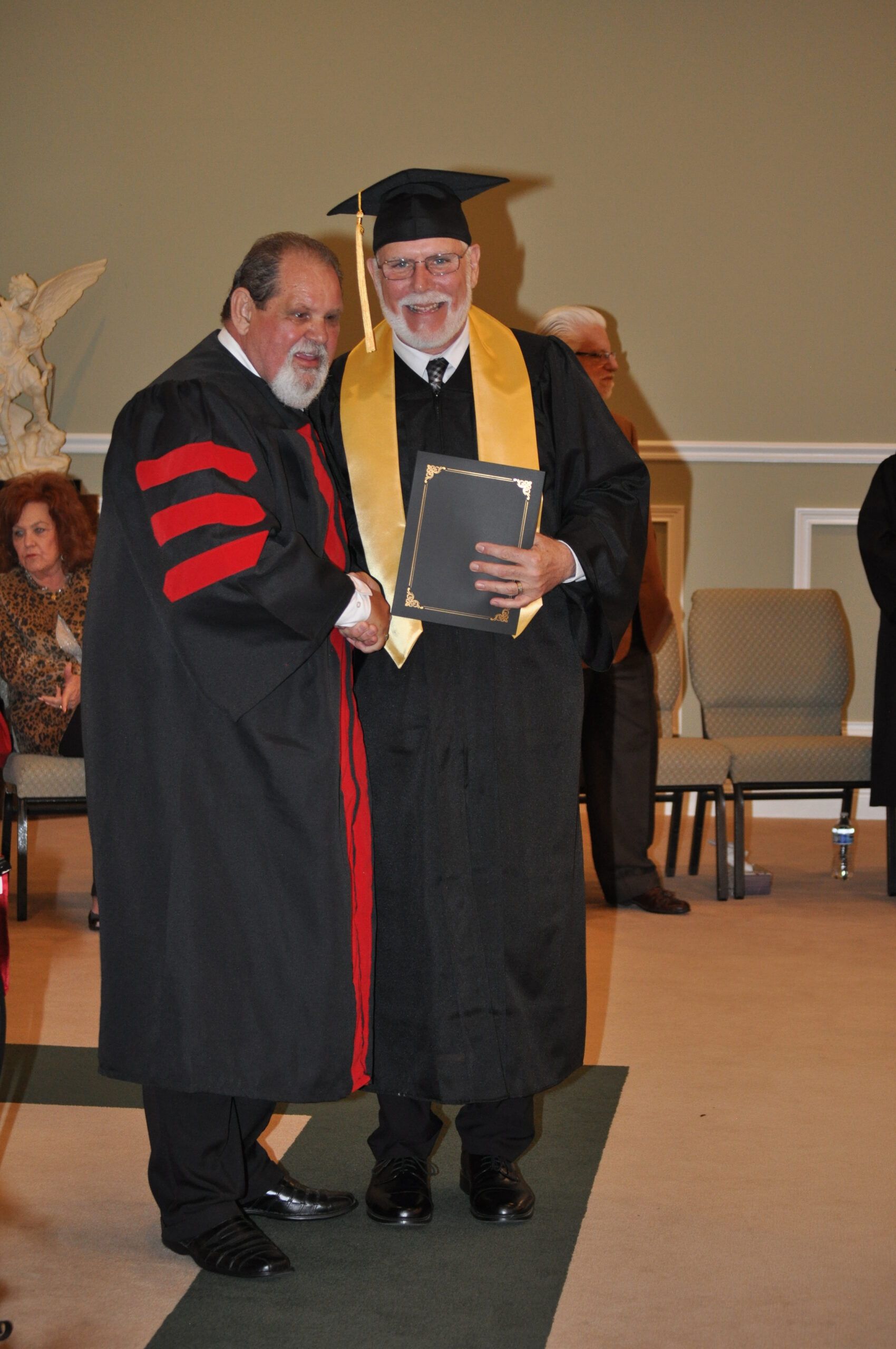 Man in graduation gown shakes hands with another man, accepting a diploma.