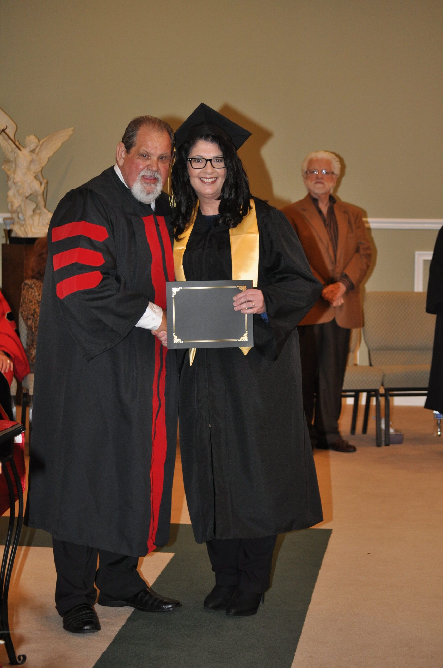 A graduate receives a diploma from a robed figure at a ceremony; a man in a brown jacket observes.
