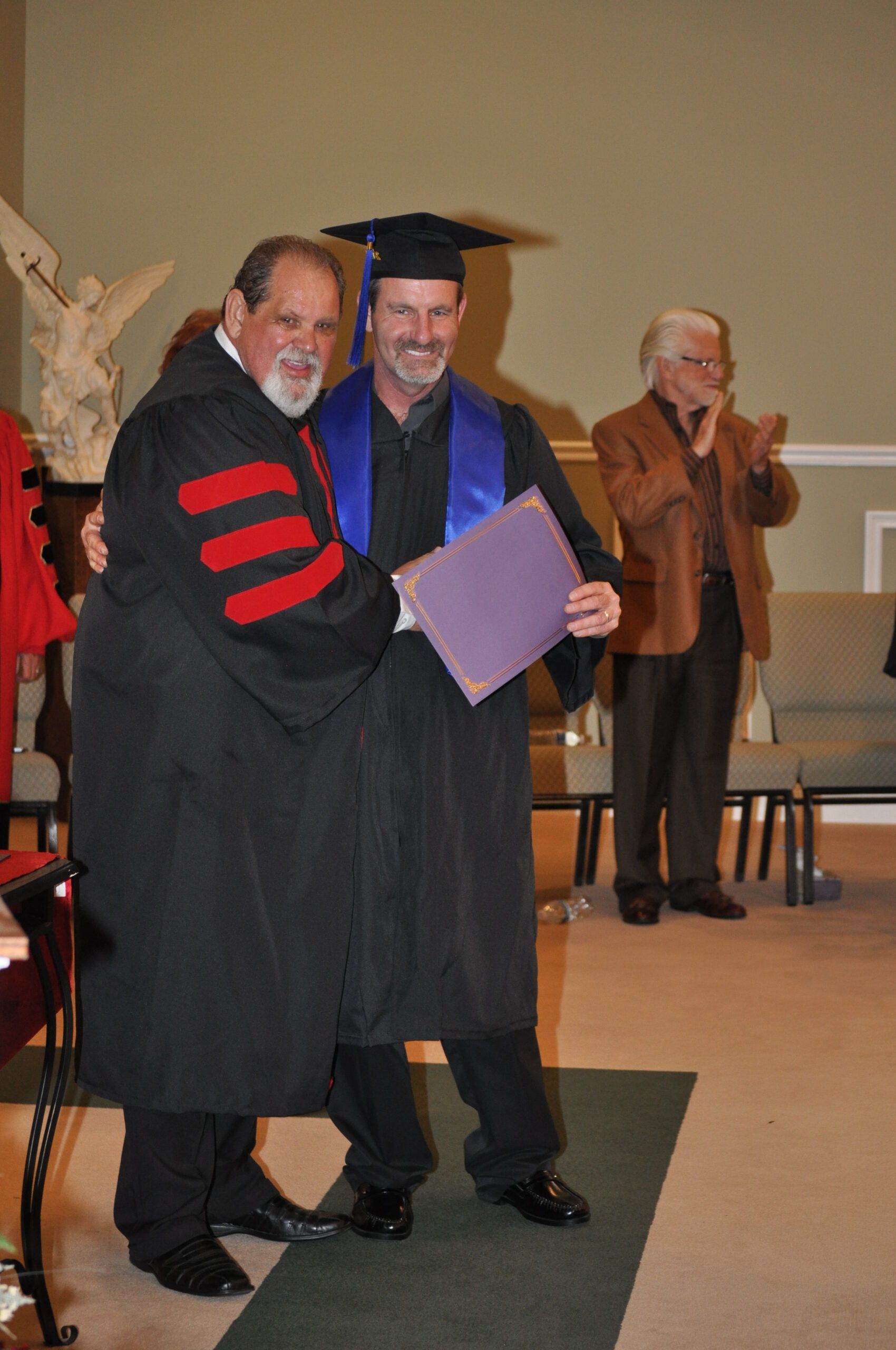 Man in graduation attire being embraced by another man in academic robes, indoors.