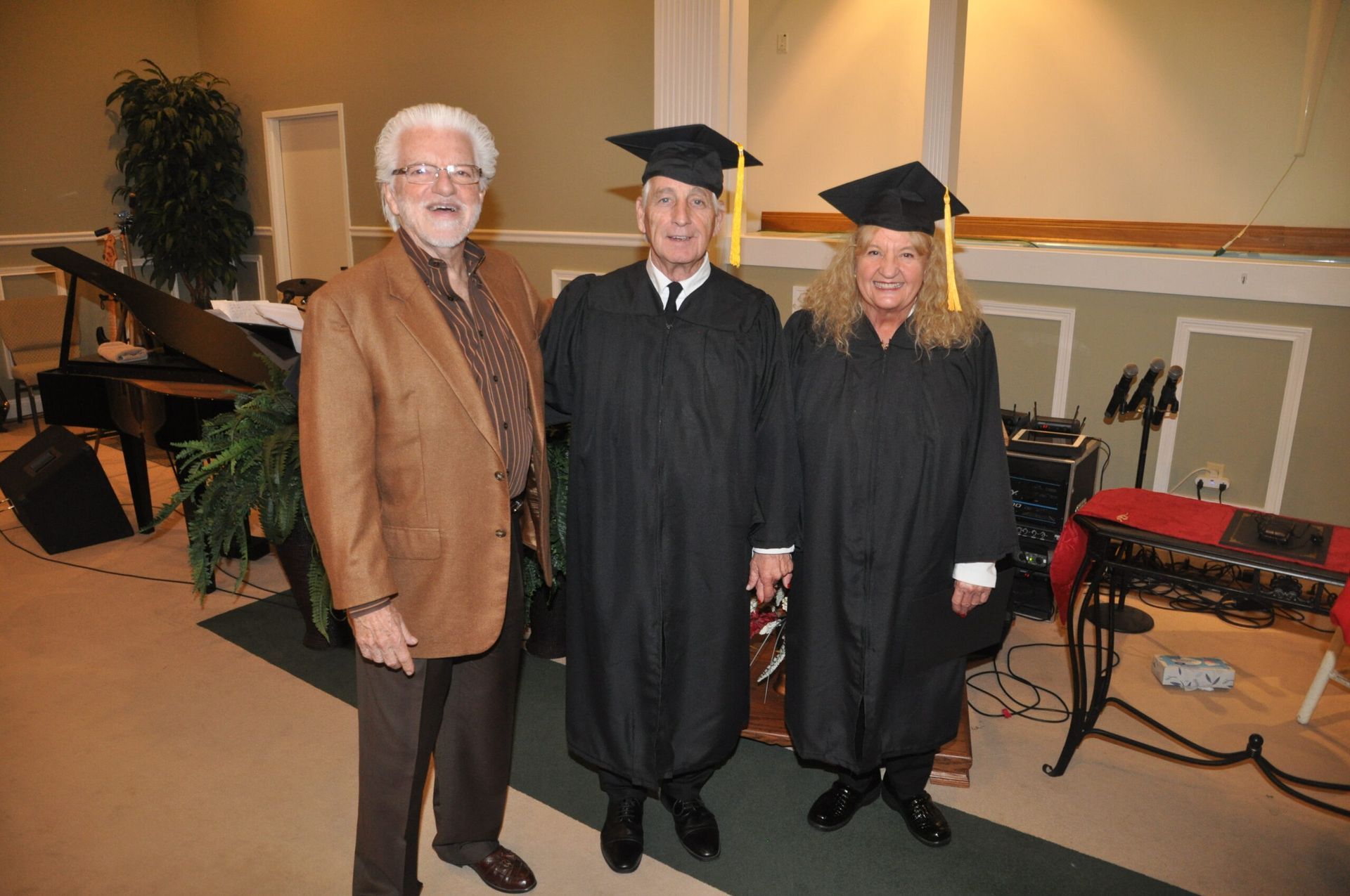 Three men, two in graduation gowns, pose inside. One man wears a brown jacket. A piano and equipment are in the background.