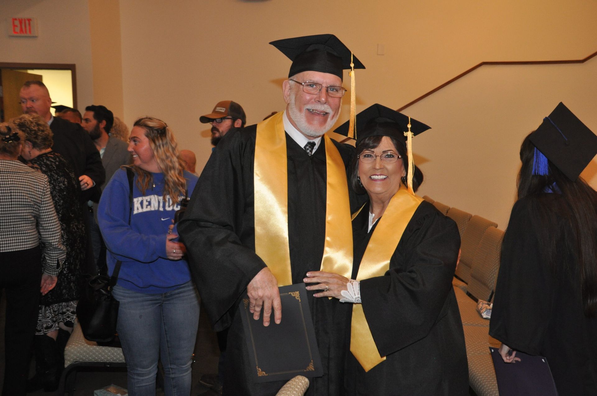 Two graduates in caps and gowns smile, holding diplomas. Gold stoles. Hallway setting.