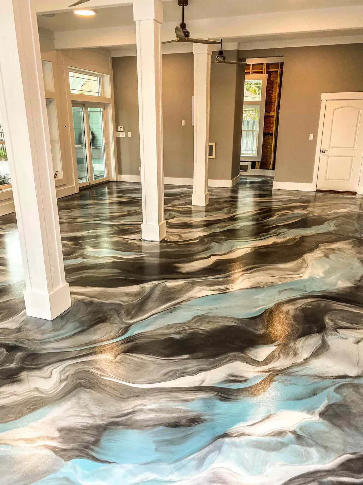 A large room with a glossy, swirled epoxy floor in shades of black, silver, blue, and white. White support columns are visible. A large room with a glossy, swirled epoxy floor in shades of black, silver, blue, and white. White support columns are visible.