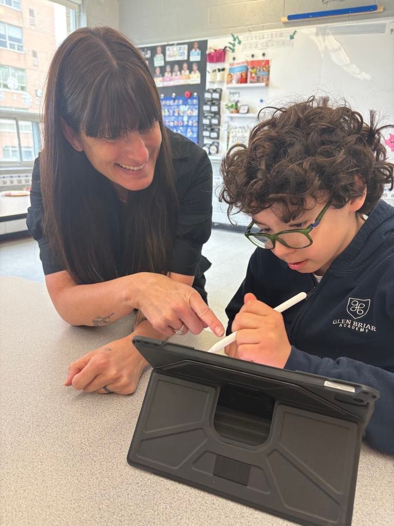 Two students work together at a table, one pointing while the other writes on a tablet.