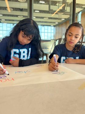 Two children writing on poster paper at a table in a bright classroom.
