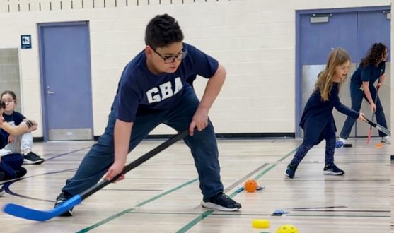 Kids playing floor hockey in a gym, using blue sticks and chasing a small orange ball.