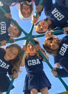 Smiling cheerleaders in blue GBA uniforms huddle in a circle, looking down at the camera against the sky.