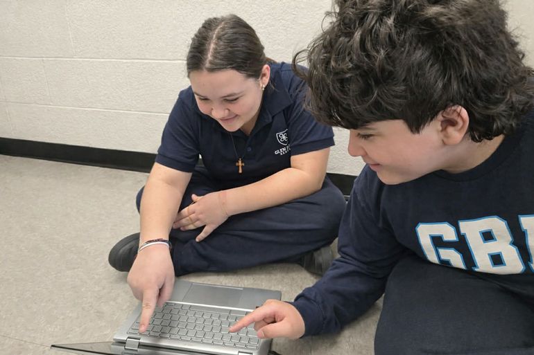 Two students kneel over a laptop on the floor, looking at the screen in a classroom.