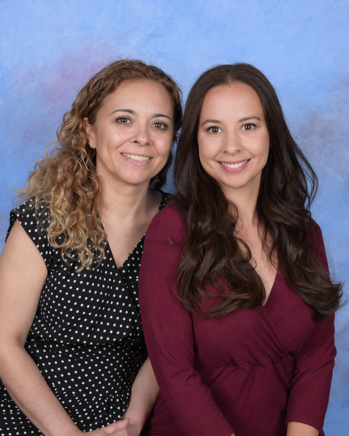 Two women smiling against a blue studio backdrop, wearing black polka-dot and burgundy tops.
