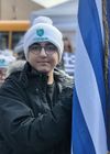 Person in a white beanie and glasses stands beside a blue-and-white flag outdoors.