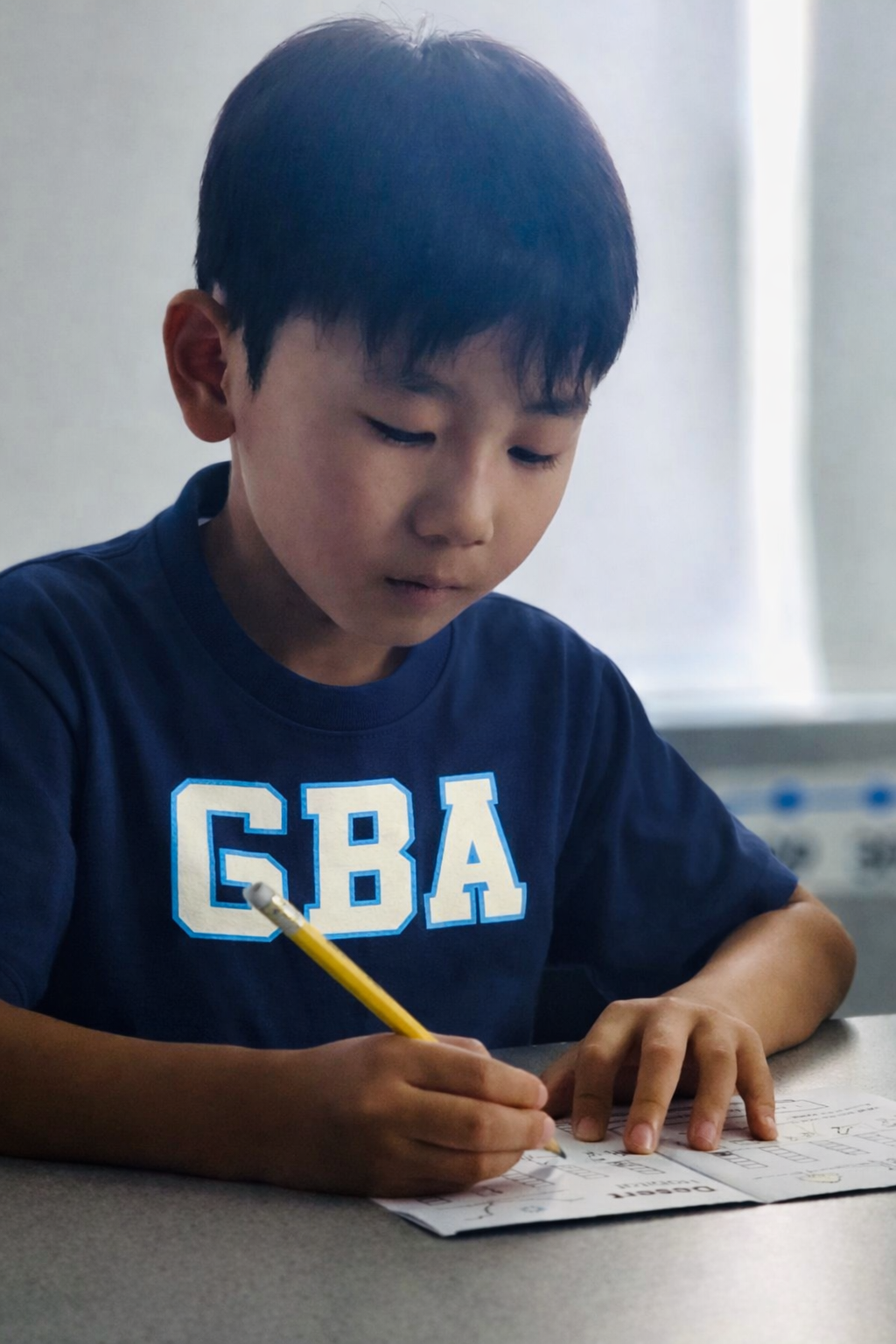 Boy in a blue GBA shirt writing with a pencil at a desk.