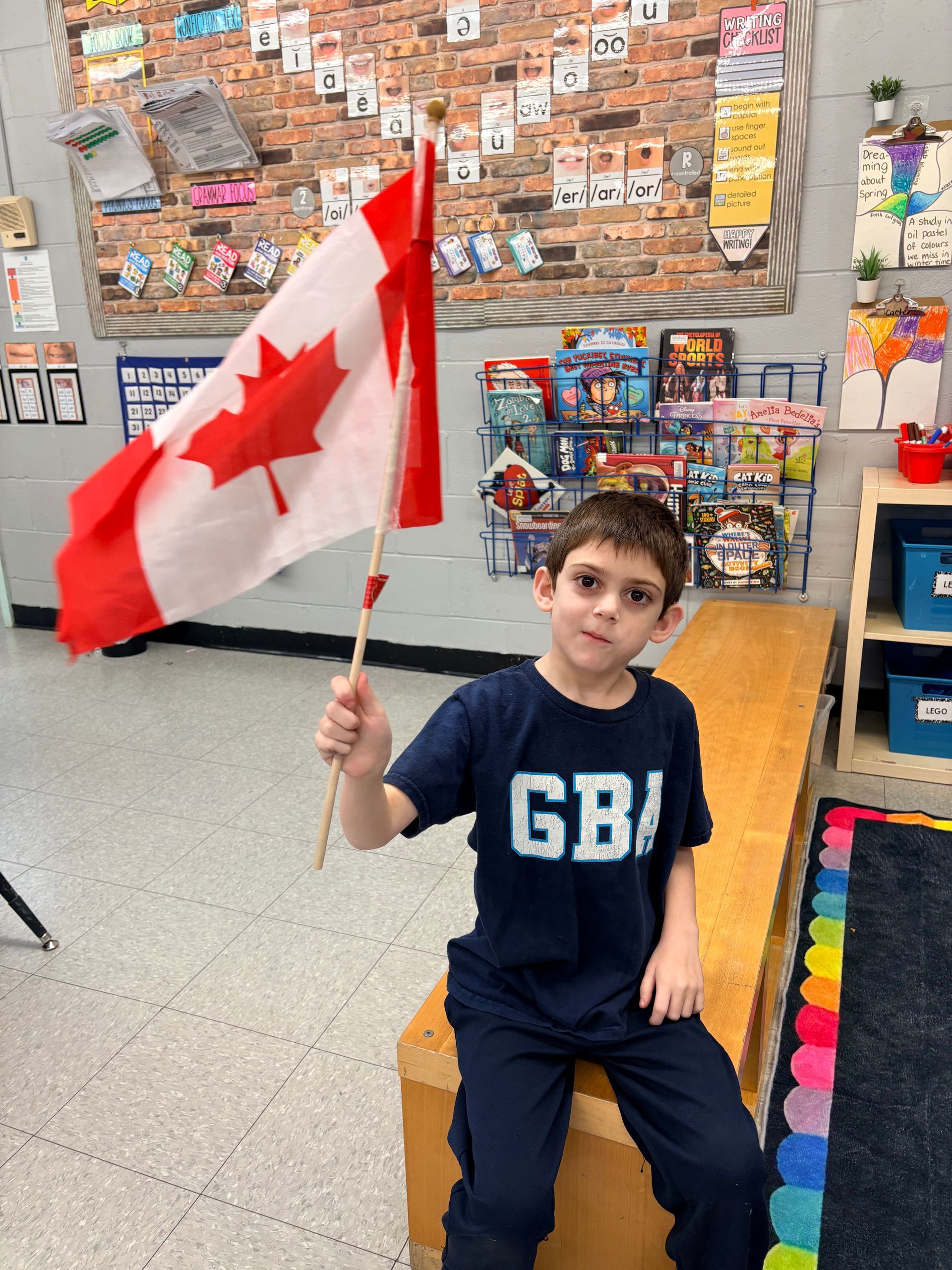 Child holding a Canadian flag in a classroom, sitting on a block with displays behind them.