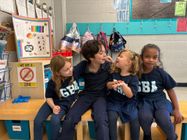 Four children in blue shirts sit together in a classroom, smiling and talking by a colorful wall display.