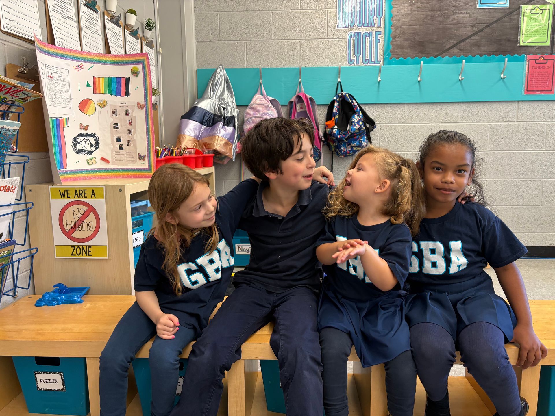 Four children in blue shirts sit together in a classroom, smiling and talking by a colorful wall display.