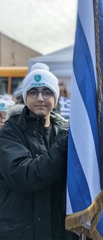 Person in winter hat and glasses holding a blue-and-white flag outdoors.