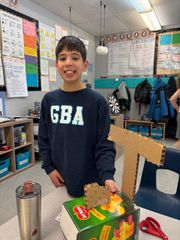 Boy standing in a classroom beside a decorated project, smiling near a table with supplies.
