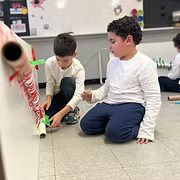 Two children on a classroom floor operating a red-and-white toy rocket launcher