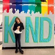 Person standing in front of a colorful “KIND” mural, holding a white object.
